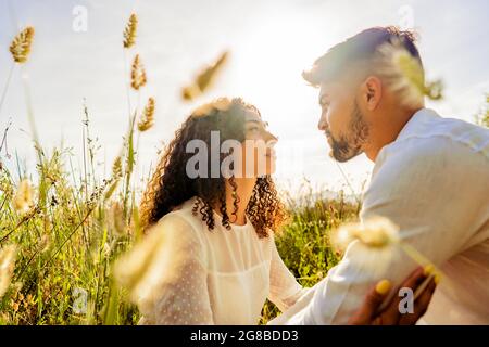 Romantische Szene von multirassischen leidenschaftlichen jungen Paar in der Liebe in die Augen einander unter hohem Gras Vegetation bei Sonnenuntergang oder Morgendämmerung mit Sonne Backli schauen Stockfoto