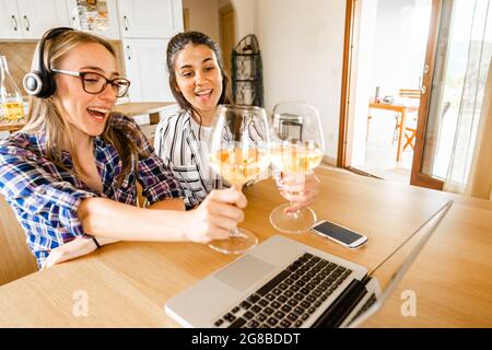 Zwei glückliche junge Frauen zu Hause, die am Tisch sitzen und einen Laptop mit Champagner oder Weißweingläsern toasten sehen. Neue normale Feier online aufgrund von soci Stockfoto