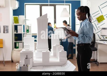 afroamerikanische Architektin Frau, die an einem Tablet arbeitet und Modellmaquette baut. Professioneller Ingenieur mit Blaupause-Planungstechnologie, der sich ein modernes Designkonzept ansehen kann Stockfoto