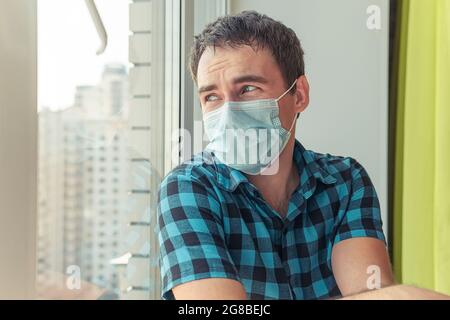 Porträt eines traurigen jungen Mannes in medizinischer Maske, der durch das Fenster aus dem Fenster schaut, beobachtet Quarantäne. Lockdown Stockfoto
