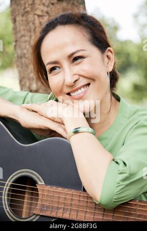 Porträt einer lächelnden, hübschen älteren Frau, die sich auf eine Akustikgitarre stützt und über etwas nachdenkt Stockfoto