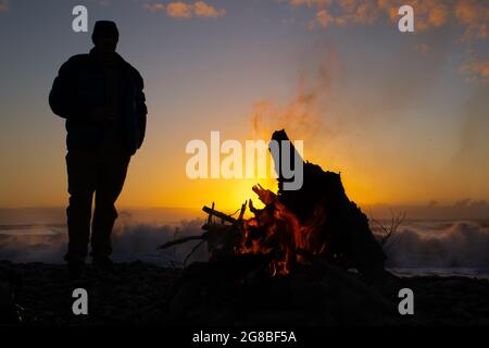 Die Sonne geht über dem Meer unter und leuchtet hinter einer männlichen Silhouette, während er sich erwärmt und in einem Winterurlaub an einem Treibholzfeuer einen Drink genießt. NZ Stockfoto