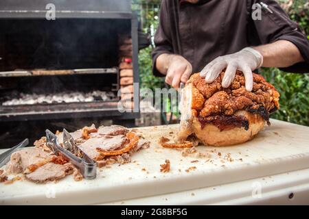 Nahaufnahme der Hände des Küchenchefs, die in einem Restaurantgarten für ein Mittagessen oder einen Brunch mit Barbecue im Hintergrund typisch italienische gebratene Schweinebacken-Porchetta schneiden Stockfoto