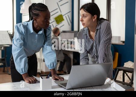 Professionelle diverse Architektinnen, die an einem Laptop-Computer an Blaupausen für den Bau von Modellmaquette arbeiten. Multiethnisches Team im Büro, das sich mit dem Entwurf eines Bauprojekts ansieht Stockfoto