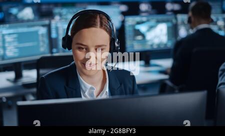 Happy Beautiful Technical Customer Support Specialist spricht an einem Headset, während er an einem Computer in einem dunklen Überwachungs- und Kontrollraum arbeitet Stockfoto