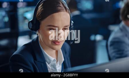 Glücklicher, selbstbewusster technischer Kundendienstmitarbeiter, der einen Headset-Anruf bei der Arbeit an einem Computer in einem dunklen Überwachungs- und Kontrollraum führt Stockfoto
