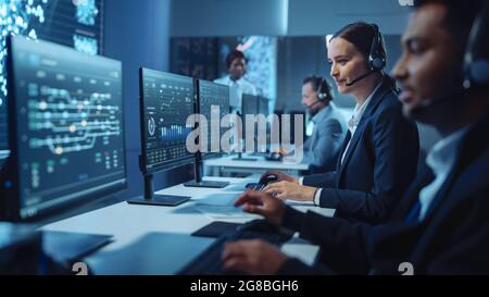 Glücklicher, selbstbewusster technischer Kundendienstmitarbeiter, der einen Headset-Anruf bei der Arbeit an einem Computer in einem dunklen Überwachungs- und Kontrollraum führt Stockfoto