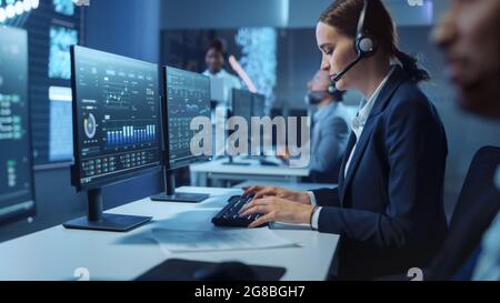 Happy Beautiful Technical Customer Support Specialist Einen Headset-Anruf während der Arbeit an einem Computer in einem dunklen Überwachungs- und Kontrollraum gefüllt Stockfoto