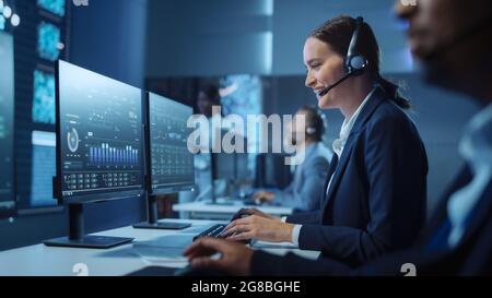 Glücklicher, selbstbewusster technischer Kundendienstmitarbeiter, der einen Headset-Anruf bei der Arbeit an einem Computer in einem dunklen Überwachungs- und Kontrollraum führt Stockfoto
