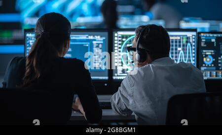 Zwei Spezialisten arbeiten an einem Computer mit Live-Ananlyse-Feed von einer globalen Karte auf einem großen digitalen Bildschirm. Die Mitarbeiter sitzen mit vor den Displays Stockfoto