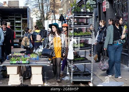 LONDON, ENGLAND - 12. Oktober 2018 Menschen kaufen Blumen auf dem Columbia Road Flower Market. Zwei hell gekleidete Mädchen gehen an Blumenvorführungen vorbei Stockfoto