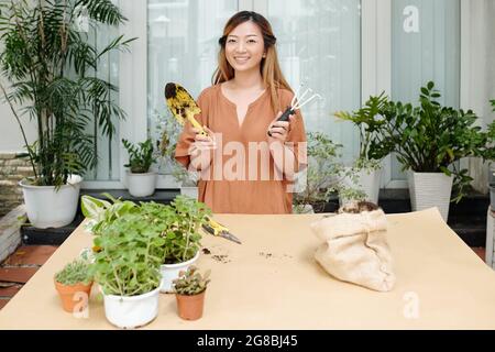 Porträt einer glücklichen jungen lächelnden Frau, die mit einer Kelle und einer Gartengabel in den Händen am großen Tisch steht und bereit ist, Blumen zu berichten Stockfoto