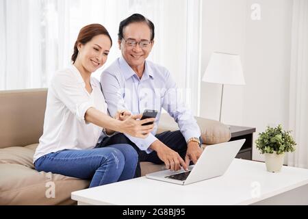 Lächelndes Seniorenpaar sitzt auf dem Sofa im Wohnzimmer und nimmt Selfie auf dem Smartphone oder mit einer mobilen Anwendung für Videoanrufe bei Familie und Freunden auf Stockfoto