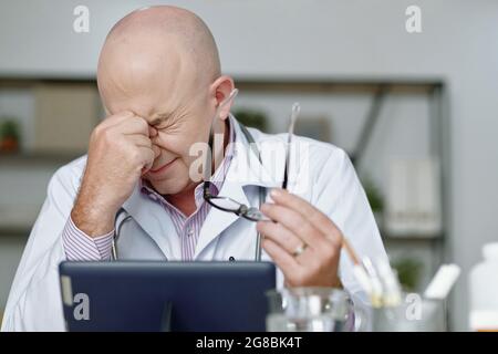 Reifer Arzt, der mit geschlossenen Augen am Tisch sitzt, sieht sehr müde aus, während er an einem digitalen Tablet arbeitet Stockfoto