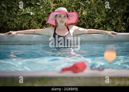 Eine asiatische Frau, die einen roten großen Hut trägt und mit einem glücklichen Gesicht im Schwimmbad steht und entspannt auf die Kamera schaut. Verfahren im Vintage-Filmstil Stockfoto