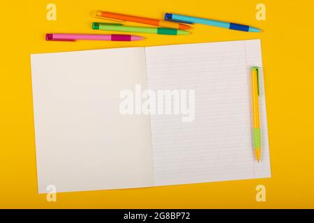 Schulnotizbuch auf gelbem Hintergrund mit Platz für Text. Leeres Blatt Papier mit schrägen Linien im Übungsheft, bunte Stifte auf einem Schreibtisch Stockfoto