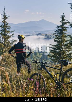 Rückansicht auf einen Radfahrer, der Uniform und Helm trägt, im Gras in der Nähe des Mountainbikes steht und die wunderschöne Berglandschaft beobachtet. Am frühen Morgen in nebligen sonnigen Bergen. Konzept der Einheit mit der Natur Stockfoto