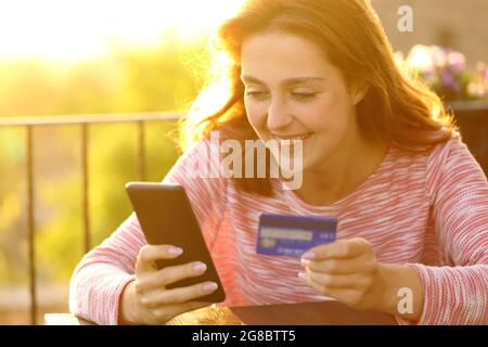 Glückliche Frau, die online mit Kreditkarte und Smartphone auf einem Balkon bei Sonnenuntergang kauft Stockfoto