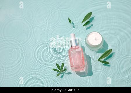 Feuchtigkeitsspendende Kosmetikprodukte mit Tropfen auf Wasser setzen. Serumglasflasche und Cremedose auf Aqua-Oberfläche mit Wellen im Sonnenlicht. Konzept für Werbung Stockfoto