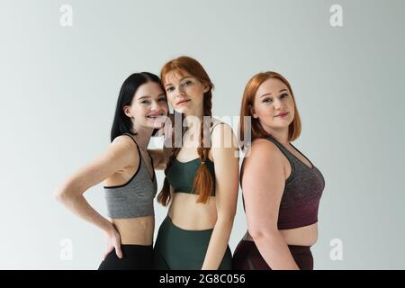 Lächelnde Sportlerin mit Vitiligo umarmenden Freund in der Nähe plus size Frau isoliert auf grau Stockfoto