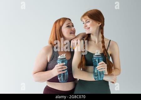 Sportlerin in Plus-Größe mit Sportflasche und lächelndem rothaarigen Freund, isoliert auf Grau Stockfoto