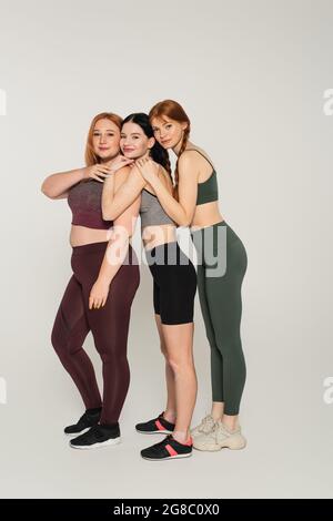In voller Länge lächelnde, körperpositive Frauen in Sportkleidung umarmen sich gegenseitig auf grauem Hintergrund Stockfoto