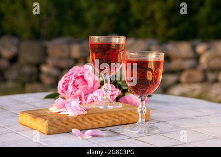 Zwei Gläser mit Rosenwein und rosa Pfingstrosen-Bouquet. Köstliches erfrischendes Getränk, riesige blühende Blumen am Tisch bei Sonnenuntergang Stockfoto