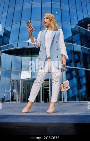 Frau im Business-Anzug steht vor einem Bürogebäude und schaut auf den Smartphone-Bildschirm. Stockfoto