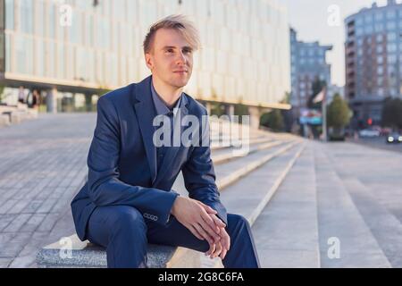 Ein junger Mann in einem Geschäftsanzug ruht vor einem Bürogebäude. Stockfoto