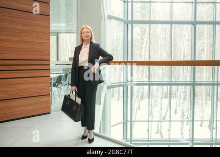Porträt einer selbstbewussten, reifen Geschäftsfrau in elegantem Anzug und mit Aktentasche, die die Kamera anschaut, während sie auf dem modernen Büroflur steht Stockfoto