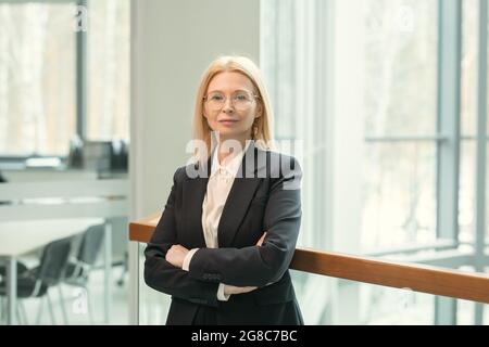 Porträt einer selbstbewussten, reifen Geschäftsfrau in Anzug, die mit gekreuzten Armen im Bürogang steht und die Kamera anschaut Stockfoto