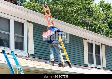 Mann, der auf einem Wohndach mit einem Paket von Schindeln zwischen zwei Leitern geht, um das Dach zu ersetzen. Stockfoto