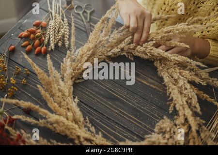 Machen rustikalen Herbst Boho Kranz. Hände halten trockenes Gras und machen stilvolle Kranz mit Wildblumen, Kräutern und Beeren auf rustikalem Holzhintergrund. H Stockfoto