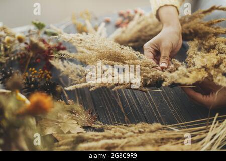 Machen rustikalen Herbst Boho Kranz. Hände halten trockenes Gras und machen stilvolle Kranz mit Wildblumen, Kräutern und Beeren auf rustikalem Holzhintergrund. H Stockfoto