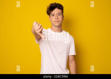 Junger Mann in einem lässigen T-Shirt, der auf einem isolierten gelben Hintergrund steht und unglücklich und wütend aussagt, Ablehnung zeigt und mit dem Daumen nach unten negativ ist Stockfoto