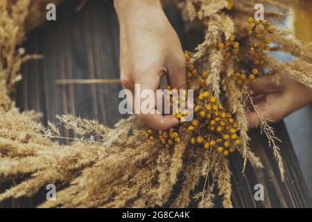 Hände, die stilvolle rustikale Herbstkranz mit trockenem Gras und duftenden Wildblumen auf dunklem Holzhintergrund machen. Ferienworkshop. Floristin in gelbem Pullover Stockfoto