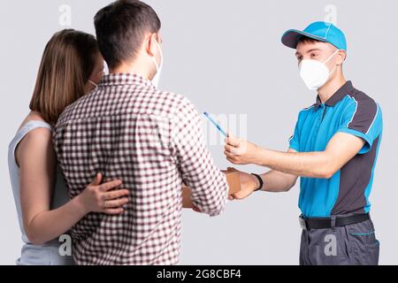 Der Kurier in einer Maske hält einen Stift aus, um die Zustellung des Paketes für das Paar in einer Maske zu bestätigen. Stockfoto