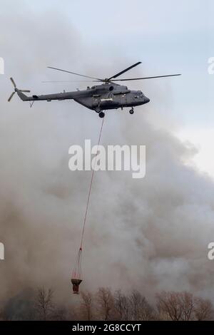 Feuerwehrhubschrauber mit einem Wasserbehälter in Rauch von einem Feuer. Bäume im Rauch eines Feuers. Stockfoto