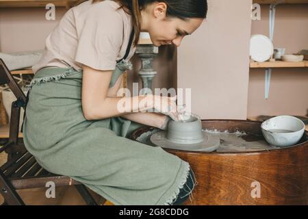Junge Frau Töpferei arbeitet in Tonstudio auf Töpferscheibe gegen Regale mit Vasen und Töpfen. Kreatives People-Konzept, Kleinunternehmen. Stockfoto
