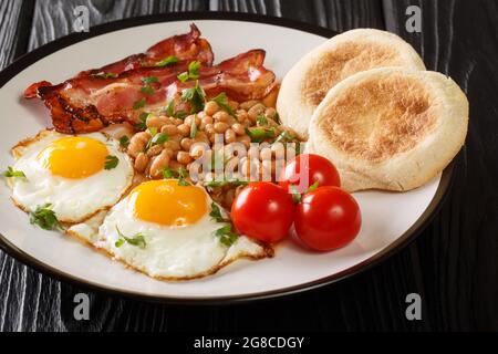 Gebratene Eier, Bohnen, Speck, Tomaten und englische Muffins in Nahaufnahme auf einem Teller auf dem Tisch. Horizontal Stockfoto
