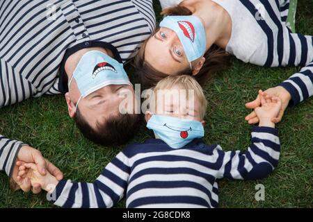 Familie - Mann, Frau und Junge in gestreiften Pullover liegen auf Gras und halten sich die Hände. Sie tragen blaue medizinische Masken mit lustigen Lächeln gemalt. Positiv, Stockfoto