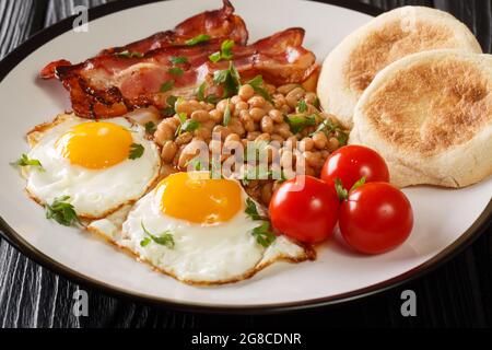 Klassisches Frühstück mit gebratenen Eiern, Bohnen, Speck, Tomaten und Muffins in der Nähe auf einem Teller auf dem Tisch. Horizontal Stockfoto