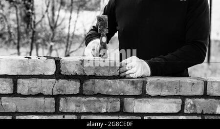 Ein Mann baut eine Mauer aus Ziegelsteinen, legt einen Ziegel auf einen Zement-Sand-Mörtel und klopft mit einem Hammer auf einen Ziegel, schwarz-weißes Foto Stockfoto