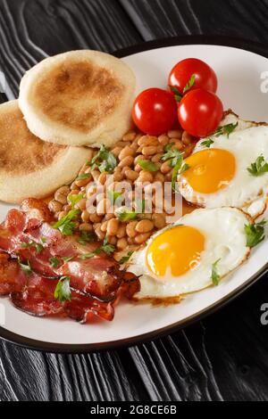 Englisches Frühstück Spiegeleier, Bohnen, Speck, Tomaten und Muffins in der Nähe auf einem Teller auf dem Tisch. Vertikal Stockfoto