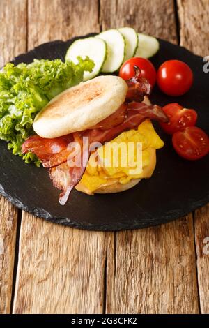 Köstliches englisches Muffin-Sandwich mit Rührei, Speck, Käse und Gemüse aus der Nähe auf einem Schieferbrett auf dem Tisch. Vertikal Stockfoto