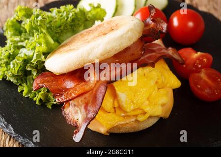 Warmes Frühstück mit englischem Muffin mit Rührei, Speck, Käse und Gemüse aus der Nähe auf einem Schieferbrett auf dem Tisch. Horizontal Stockfoto