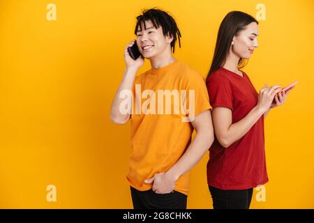 Junges multirassisches Paar, das lächelt, während es Handys benutzt und spricht, die über der gelben Wand isoliert sind Stockfoto