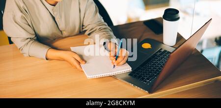 Eine Frau mit Brille studiert online mit ihrem Laptop und schreibt auf einem Notizblock. Einfache Online-Bildung mit Computer- und eLearning-Webinaren Stockfoto