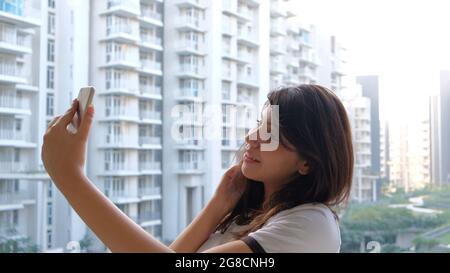 Junge indische Frau, die ein Selfie auf einem Balkon auf dem Hintergrund von Hochhäusern macht Stockfoto