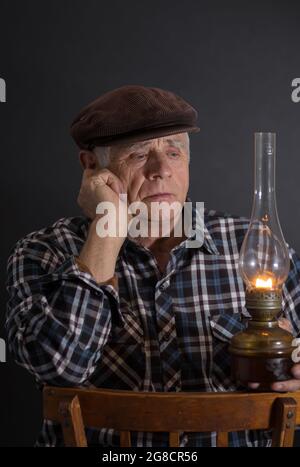 Dezernes Porträt eines älteren ukrainischen Mannes, der in der Dunkelheit eine alte Kerosin-Lampe nimmt und auf einem Holzstuhl sitzt und denkt Stockfoto